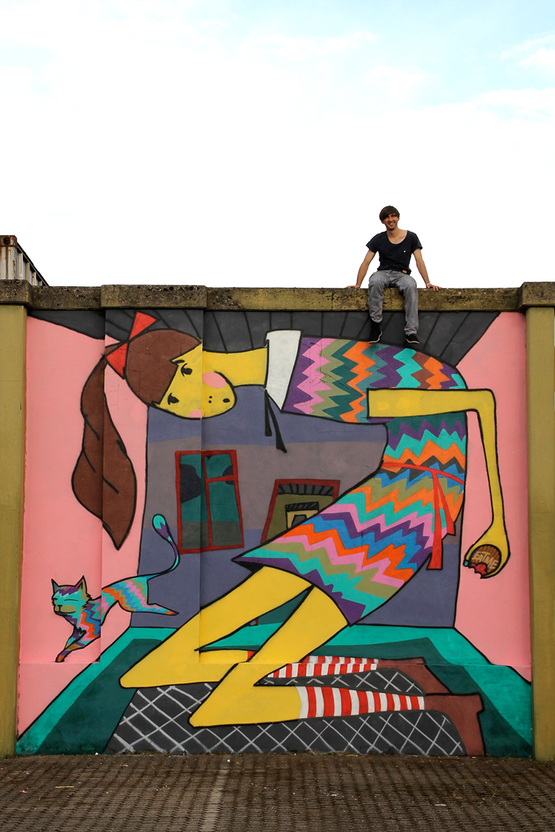 GRAFFITI-SPRAYER KÖLN-EHRENFELD: Illustration von Alice im Wunderland im Format 4m x 5m im Luisenviertel. Alice hat ein bunt gestreiftes Kleid an und eine rote Schleife im Haar. Sie ist am wachsen und stößt bereits mit dem Rücken gegen die Decke des farbenfrohen Zimmers an. Neben Alice springt ein bunte Katze. Der Graffiti-Künstler sitzt auf der bemalten Mauer.