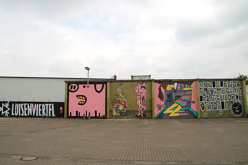 GRAFFITI-SPRAYER KÖLN-EHRENFELD: Große Außenwand mit dem Schriftzug "Luisenviertel", einem pinken Schwein einem Turm aus gestapelten Gegenständen mit einer Schere an der Spitze, einer Räuberleiter aus pinken Figuren, Alice im Wunderland, die aus einem Raum wächst, und einem Zitat mit Uhren in schwarz-weiß 