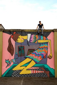 Illustration von Alice im Wunderland im Format 4m x 5m. Alice hat ein bunt gestreiftes Kleid an und eine rote Schleife im Haar. Sie ist am wachsen und stößt bereits mit dem Rücken gegen die Decke des farbenfrohen Zimmers an.Neben Alice springt ein bunte Katze. Der Grffiti-Künstler sitzt auf der bemalten Mauer.