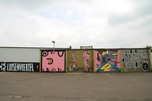 Große Außenwand mit dem Schriftzug "Luisenviertel", einem pinken Schwein einem Turm aus gestapelten Gegenständen mit einer Schere an der Spitze, einer Räuberleiter aus pinken Figuren, Alice im Wunderland, die aus einem Raum wächst, und einem Zitat mit Uhren in schwarz-weiß